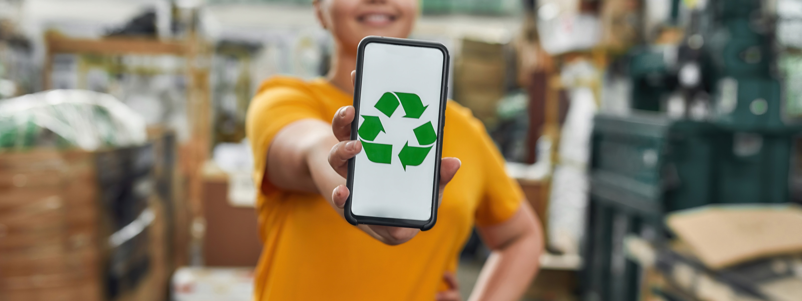 Nahaufnahme von einem Smartphone mit grünem Recyling Icon, das von einer Person mit orangem T-Shirt gehalten wird