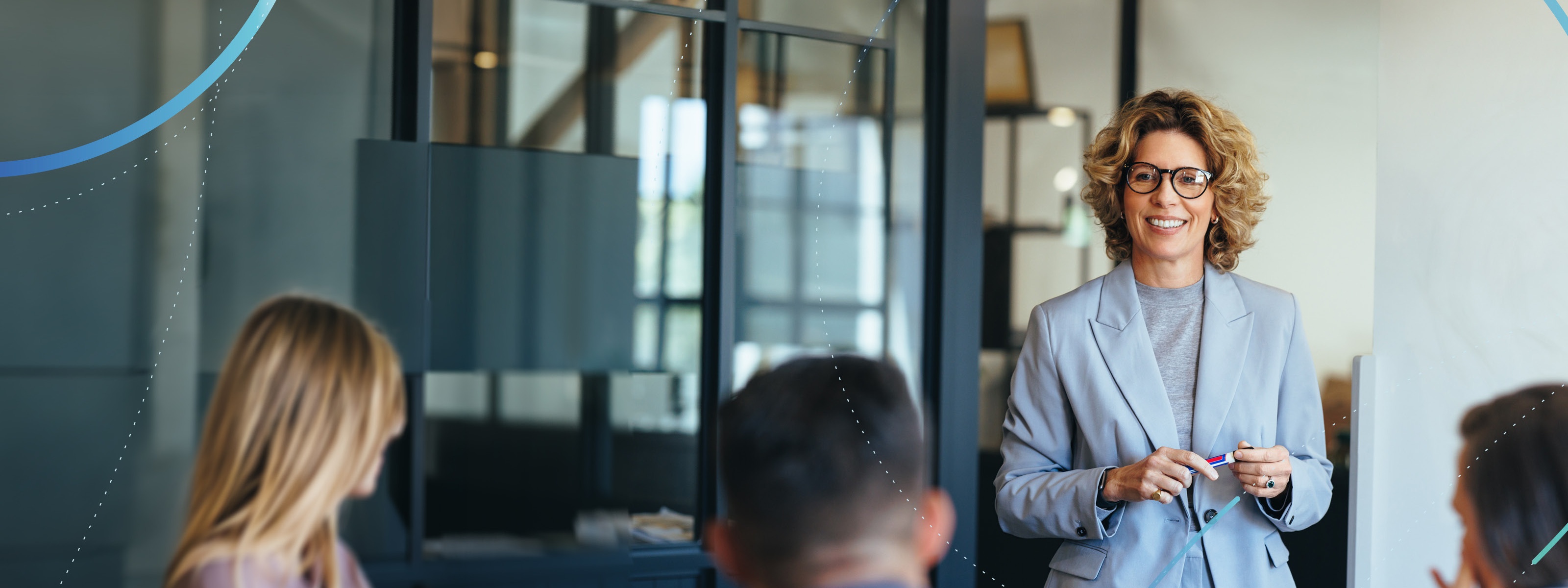Eine Person im hellen Anzug mit Brille steht lächelnd vor einer kleinen Gruppe  in einem modernen Besprechungsraum und hält einen Stift in der Hand, als würde sie etwas erklären.