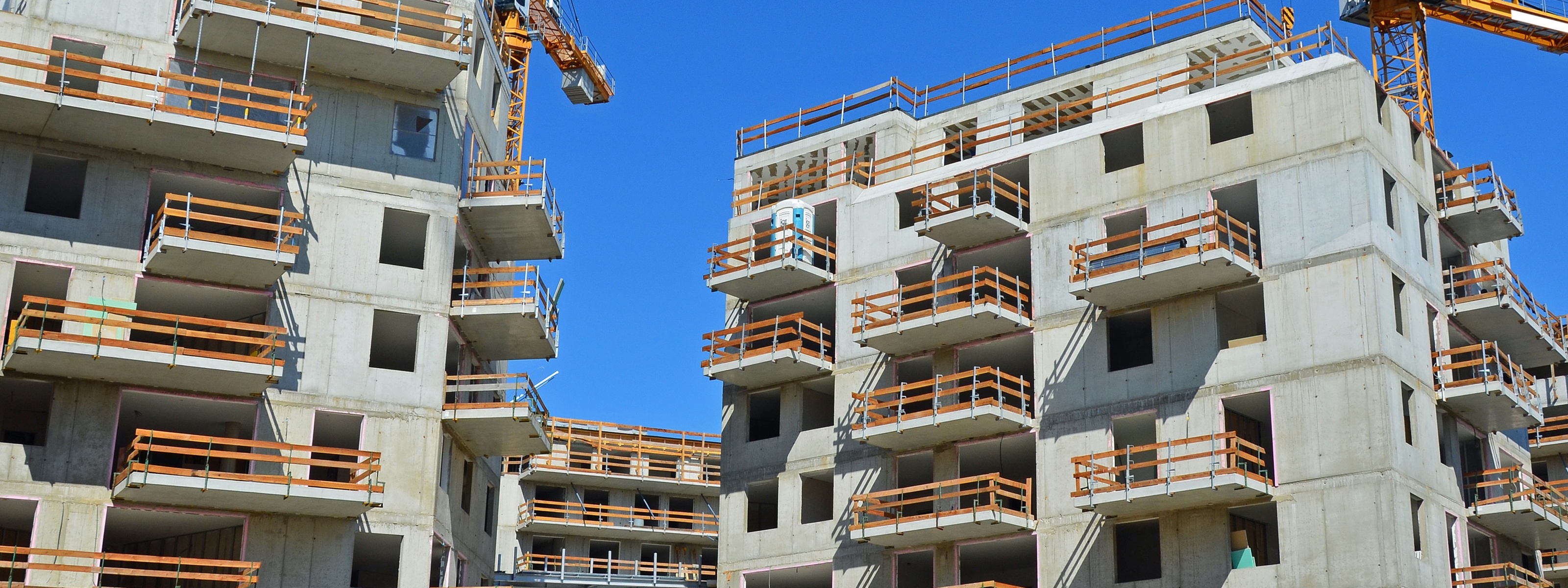 Mehrere Wohnhäuser in Bauphase mit Balkonen, im Hintergrund Kräne unter blauem Himmel