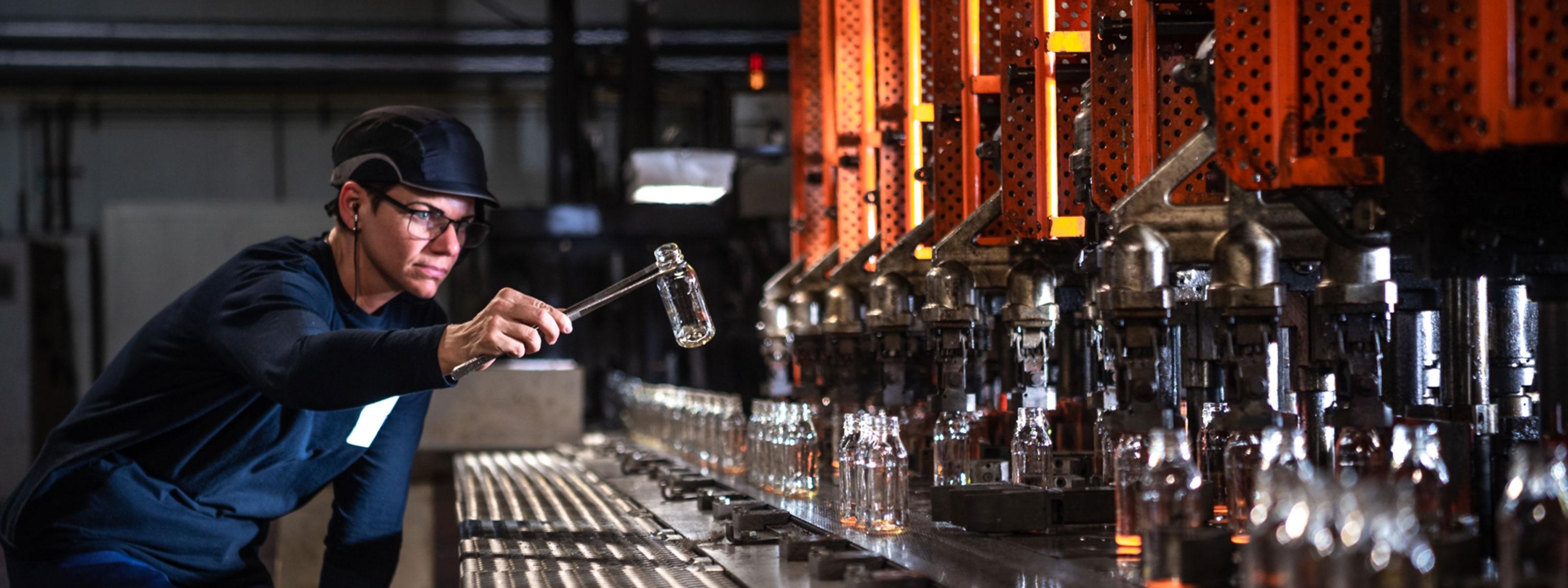 Person mit Brille und Haube vor Fließband mit Glasflaschen stehend greift mit Zange nach Glas