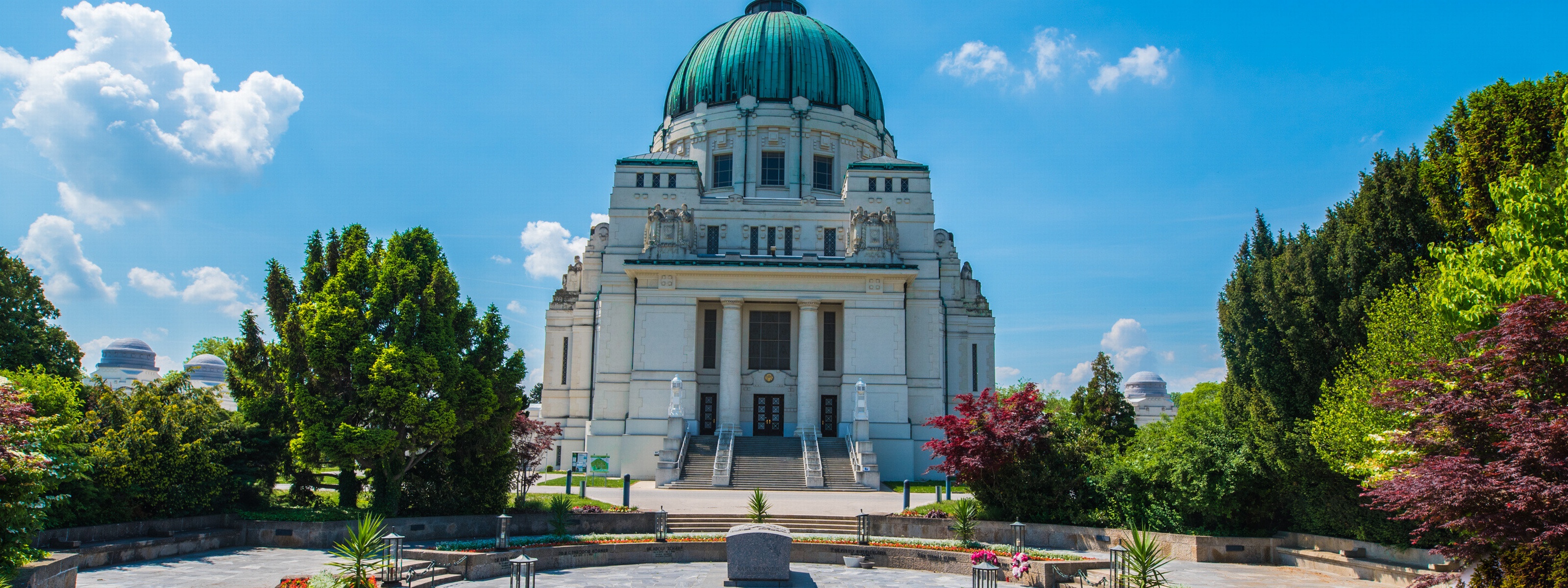 In der Mitte ist ein runder Platz mit einem steinern Sarg. Links und rechts davon sind grüne Büsche und Bäume. Neben dem Platz ist eine Aufbewahrungshalle mit einer grünen Kuppel