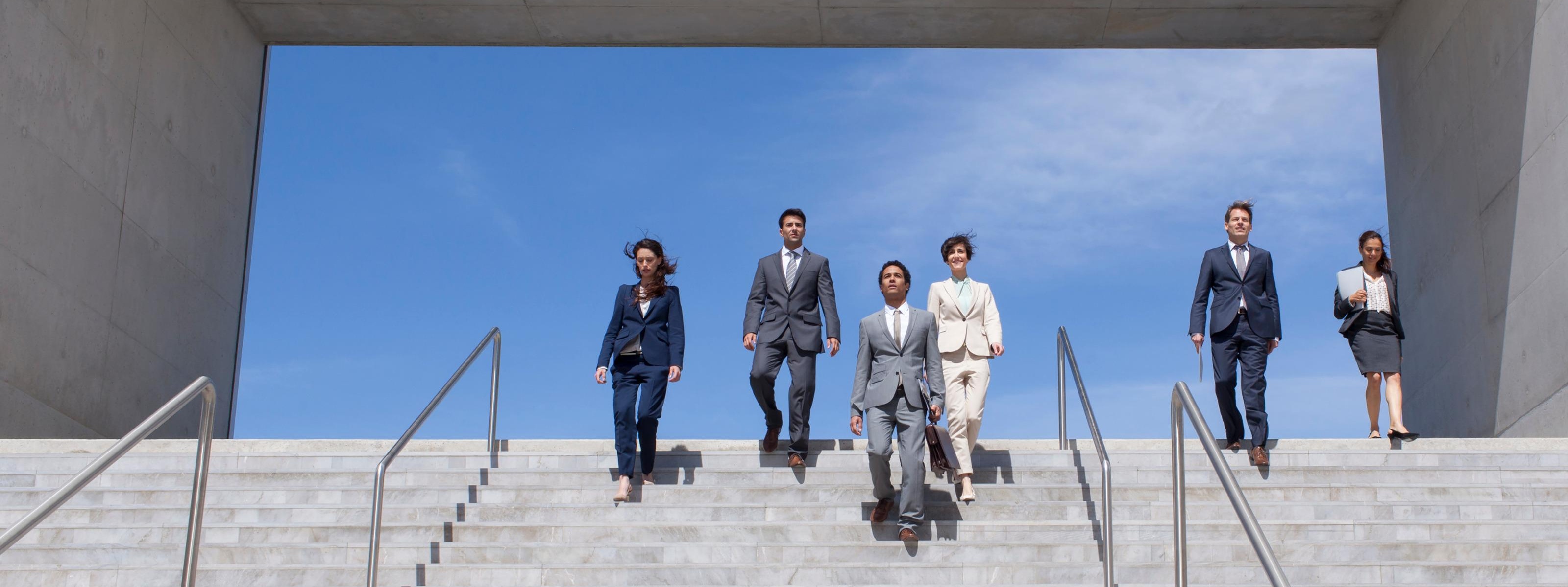 Mehrere Personen in Businesskleidung eine Betontreppe herabsteigend, im  Hintergrund blauer Himmel