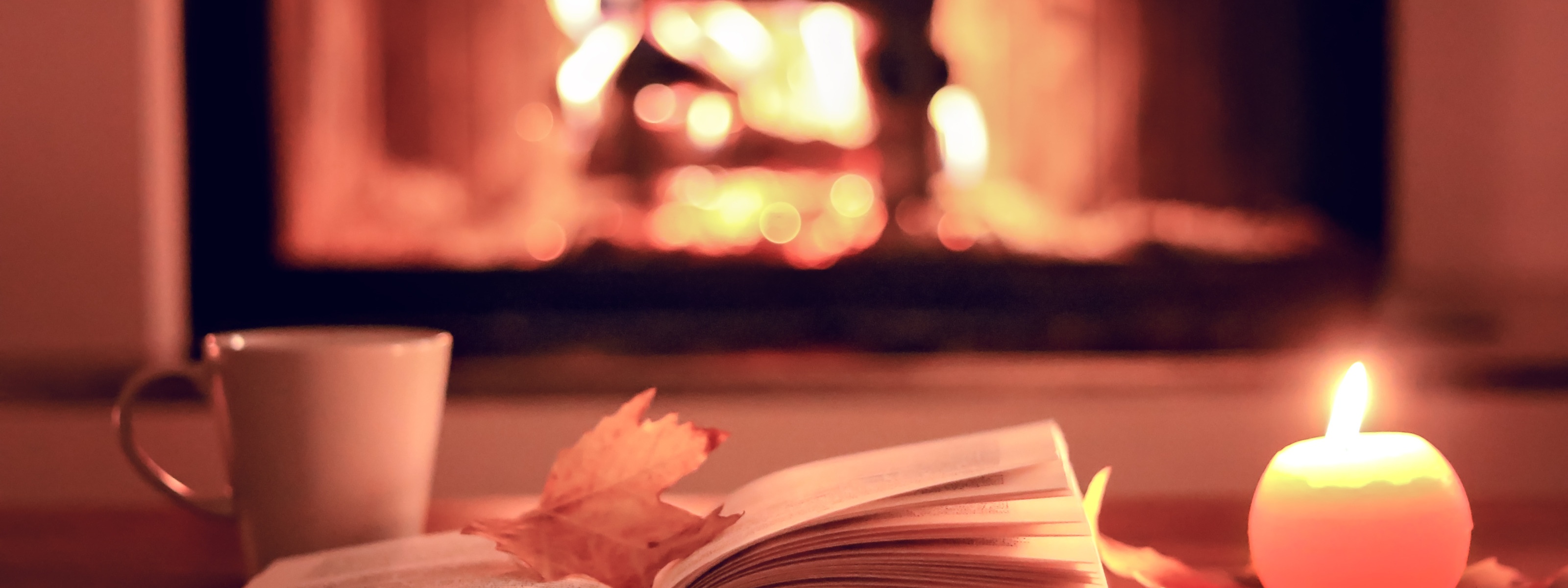 Aufgeklapptes Buch mit gelb gefärbtem herbstlichen Blatt darauf liegend, neben brennender runder Kerze und Tasse, im Hintergrund Kaminfeuer