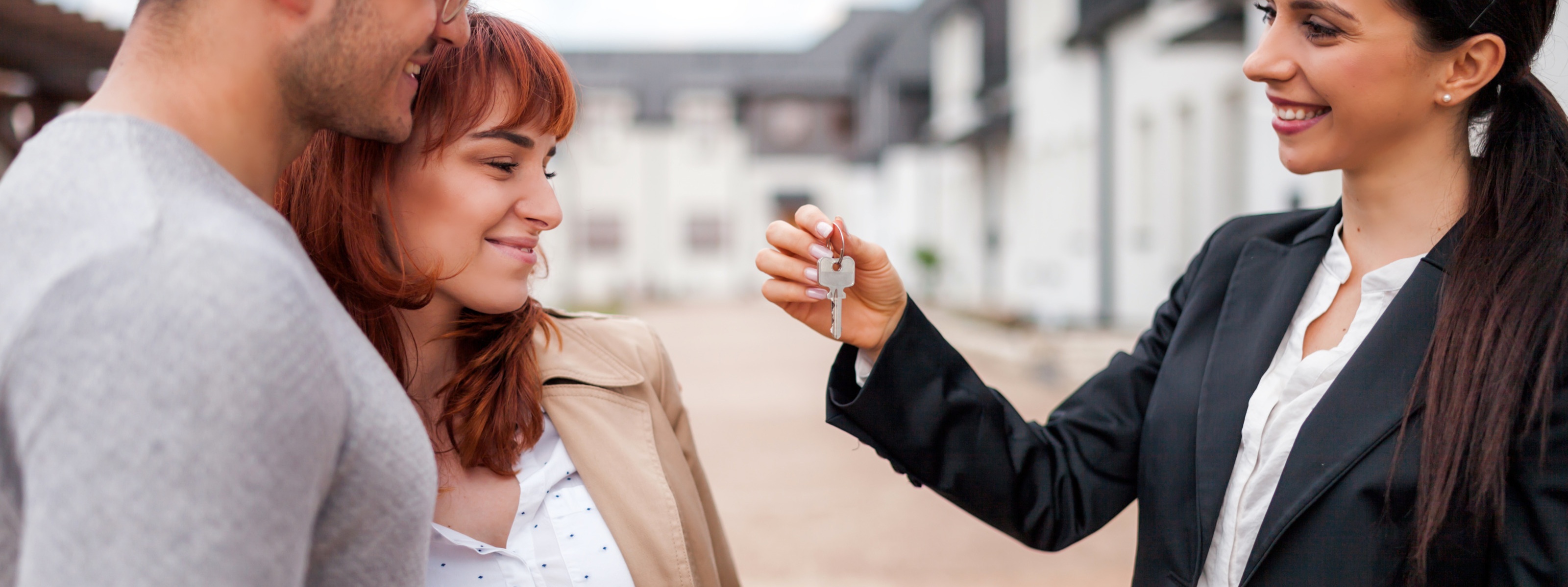 Lächelnde Person reicht zwei aneinander geschmiegten ebenfalls lächelnden Personen Schlüssel, im Hintergrund verschwommen Häuserreihen