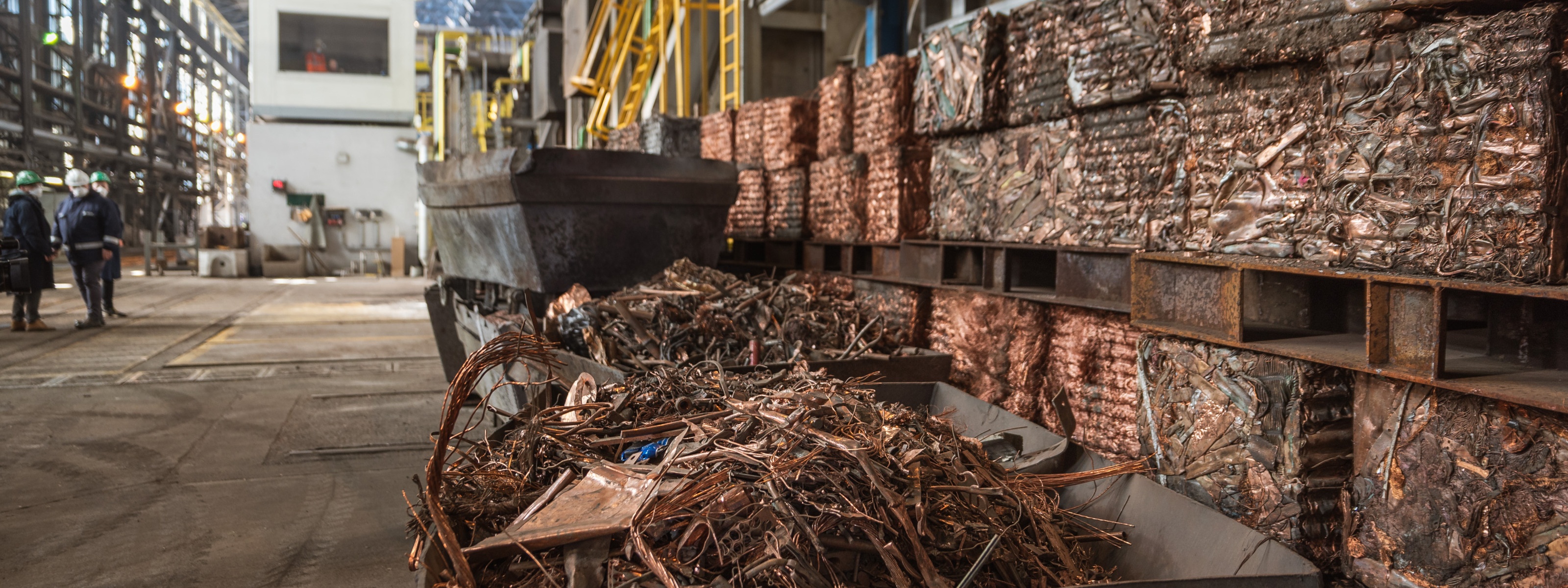 Kupferschrott in einem Container bereit für Recycling in einer Fabrikhalle, im Hintergrund stehen drei Personen in Schutzbekleidungen