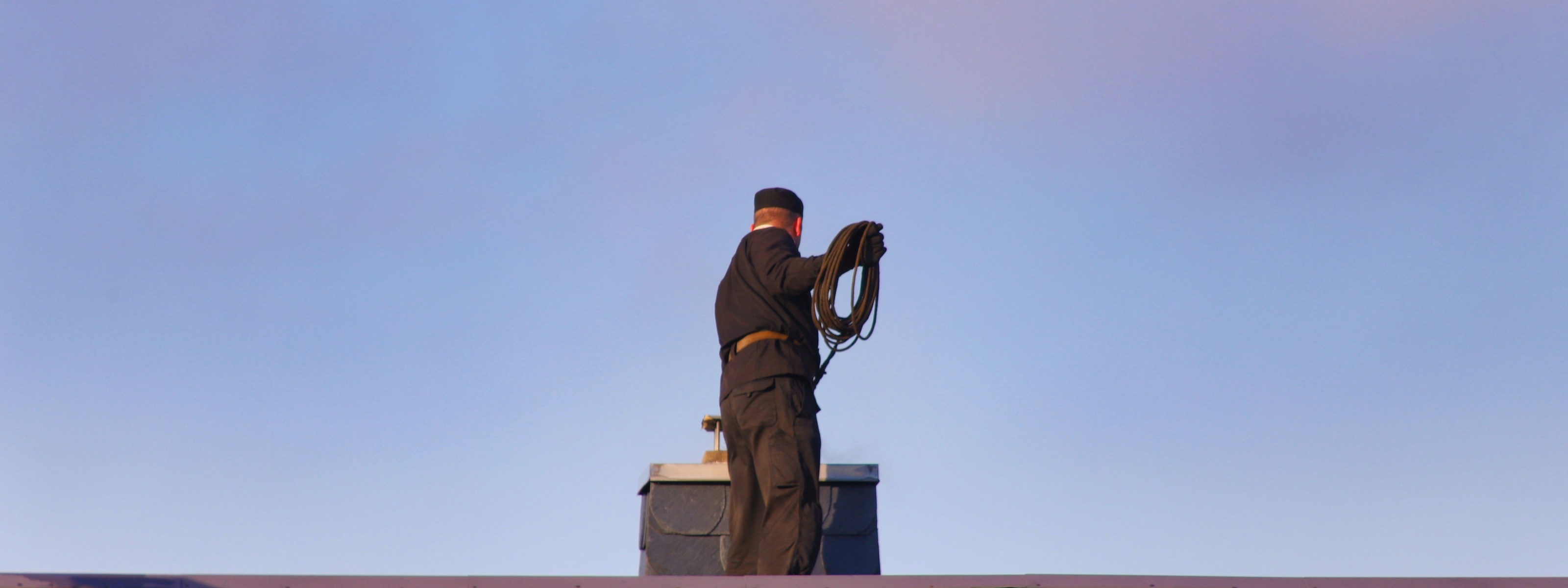 Person in Rückenansicht in schwarzer Arbeitskleidung mit schwarzer Kappe steht auf Dach vor Schornstein unter blauem Himmel und hält aufgewickeltes Drahtseil in Händen