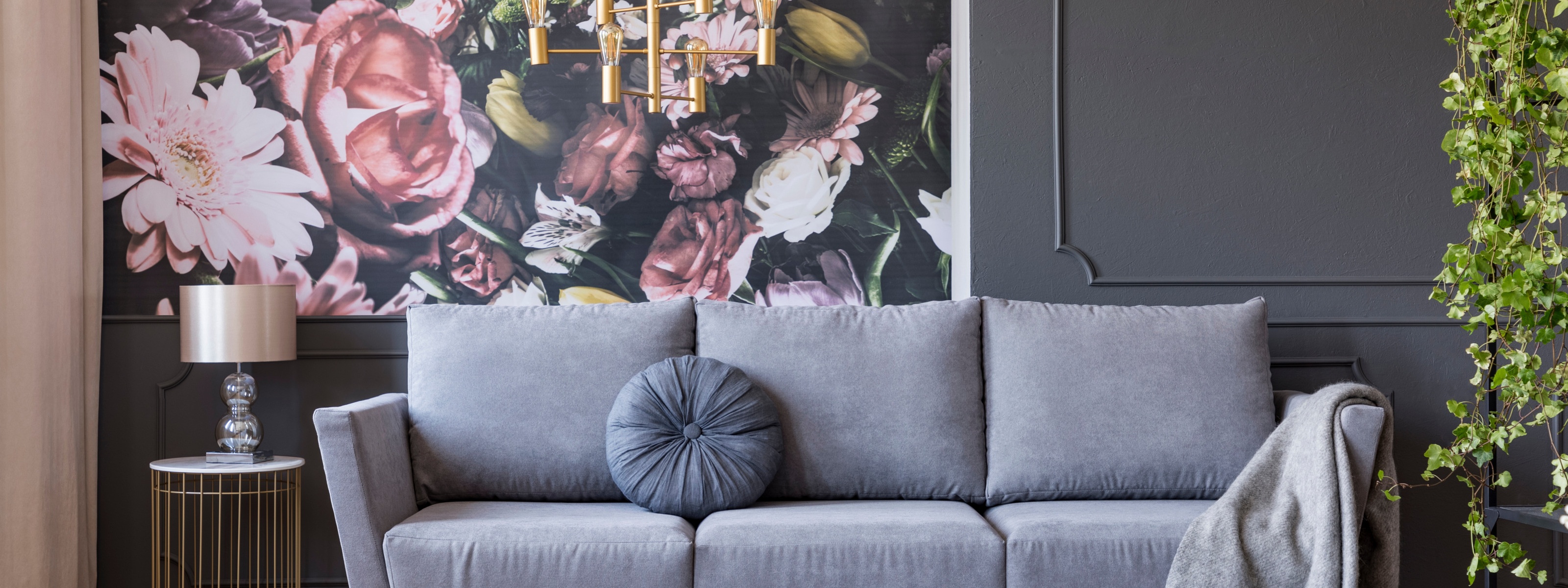 Graues Sofa vor großer Blumentapete, ringsum verschiedene Einrichtungsgegenstände