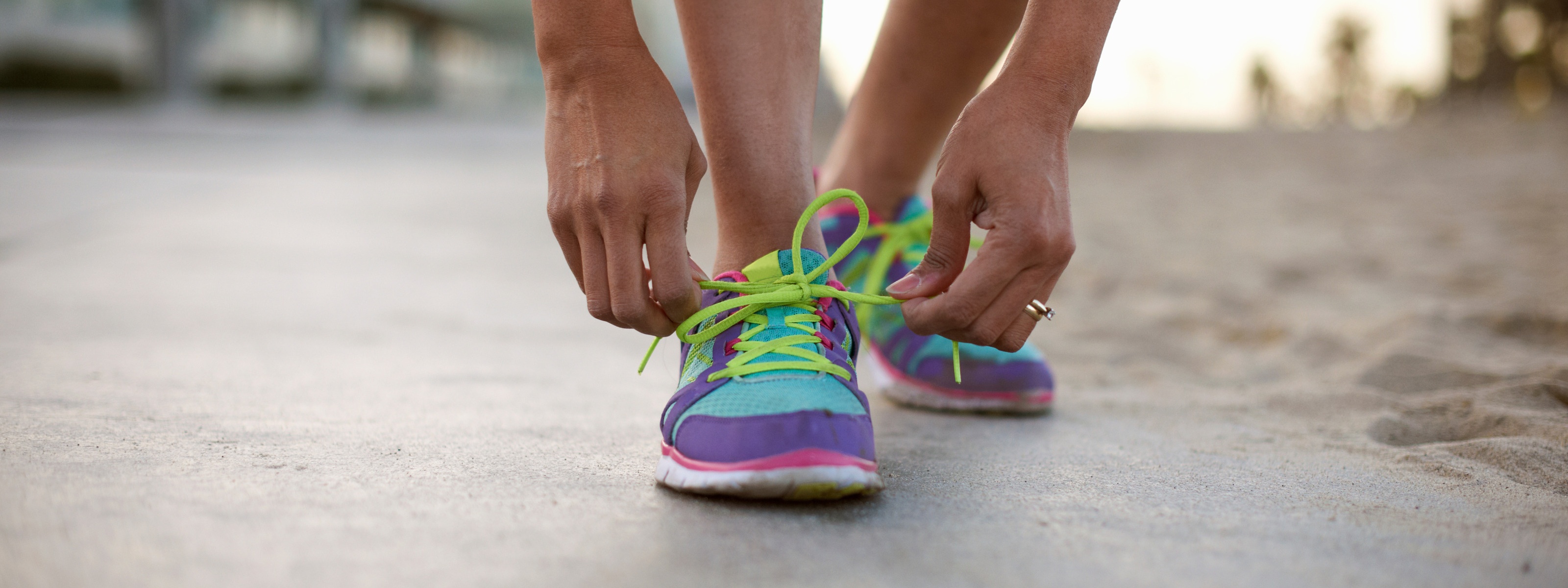 Detailansicht auf bunten Sportschuh einer Person, die sich gerade die Schnürsenkel zu Masche bindet, ringsum verschwommen Sand und Strandumgebung