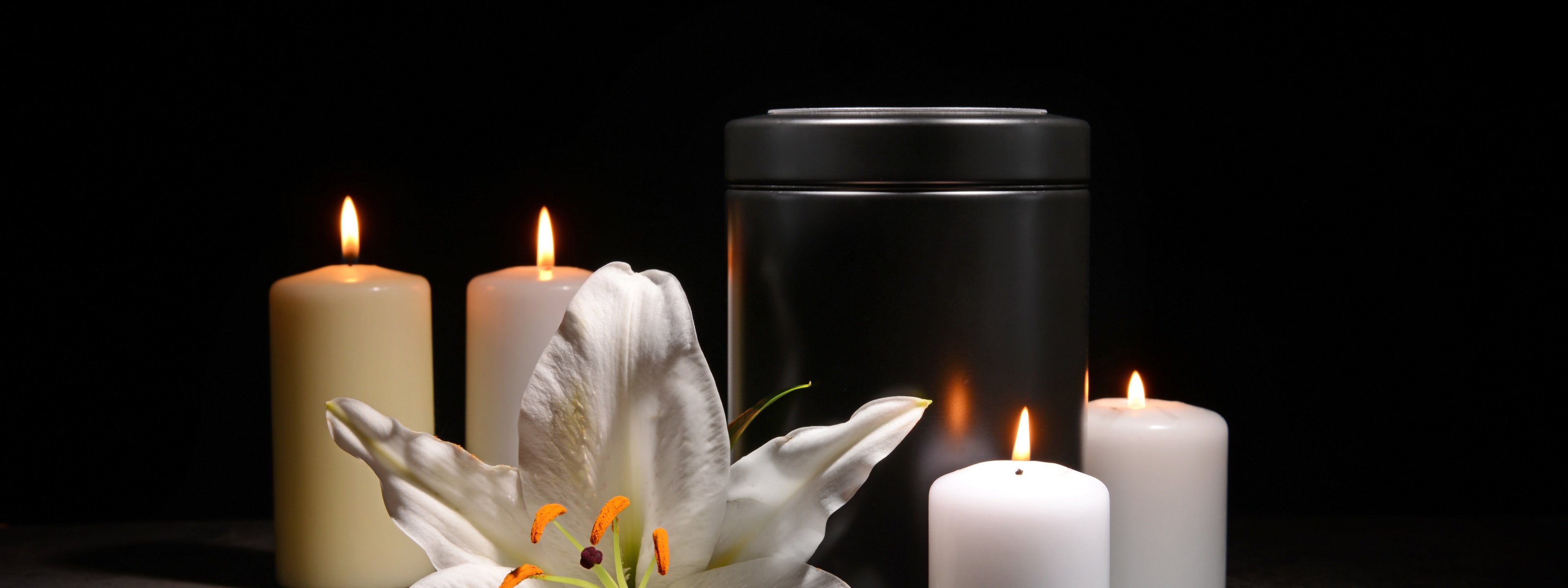 Schwarze Urne umrandest von vier weißen brennenden Kerzen und Blüte einer weißen Lilie