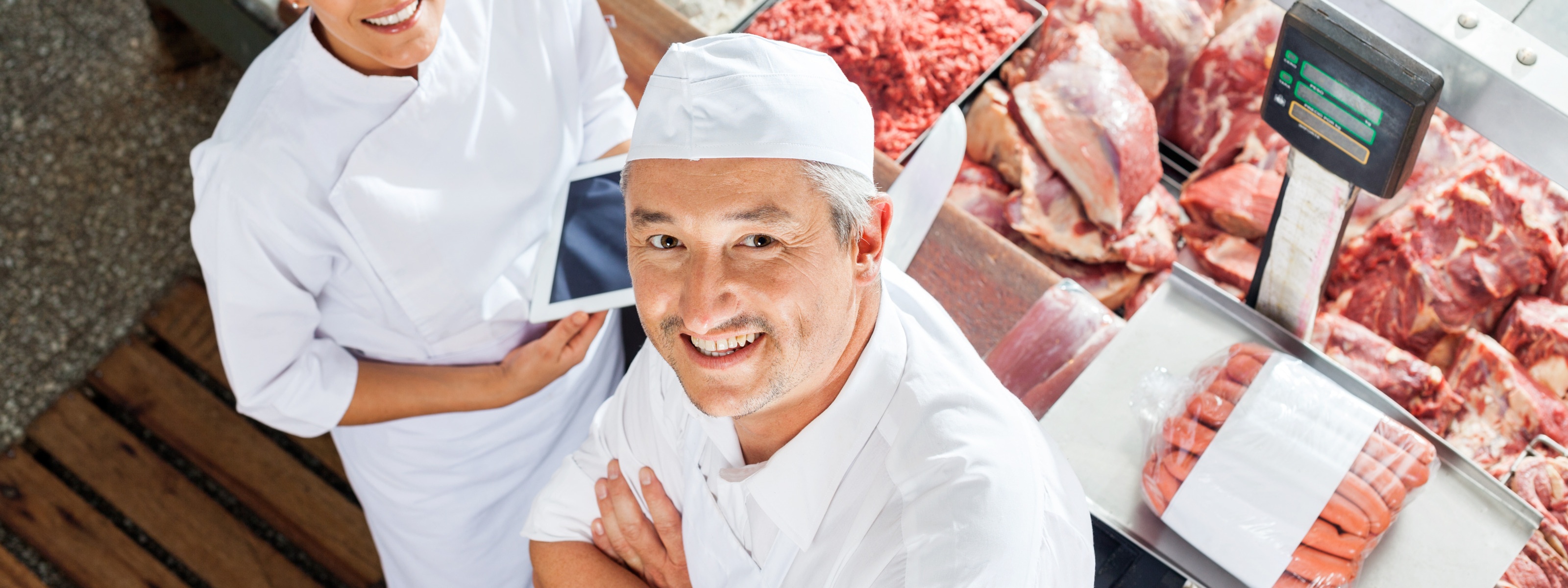 Zwei lächelnde Personen in weißem Arbeitsgewand nach oben blickend, daneben Frischetheke mit Fleischwaren