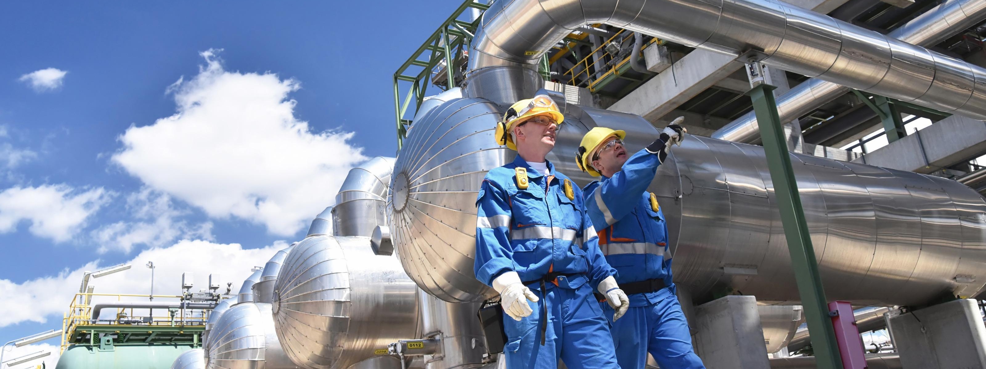Zwei Personen in Schutzausrüstung mit gelben Helmen gehen entlang Raffinerietanks, im Hintergrund blauer Himmel mit Wolken