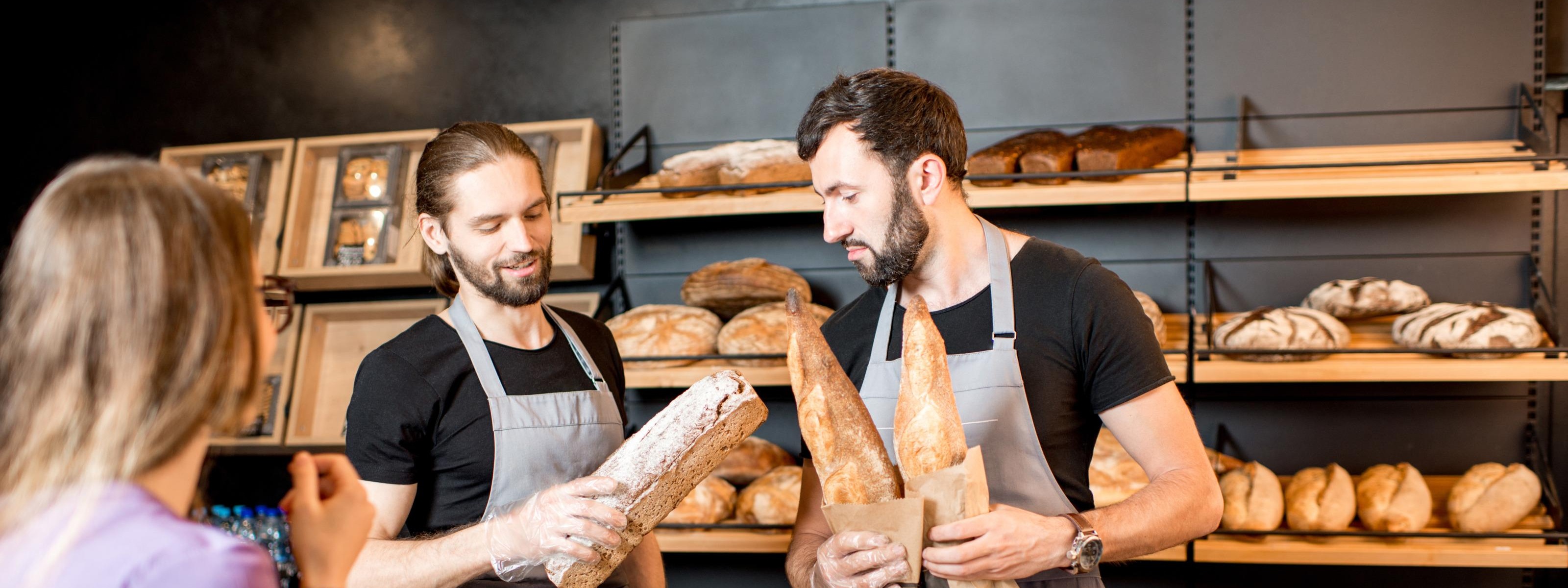 Zwei Personen mit grauen Schürzen auf Brote blickend hinter Theke stehend, eine Person im Vordergrund, im Hintergrund Regale mit Brotlaiben