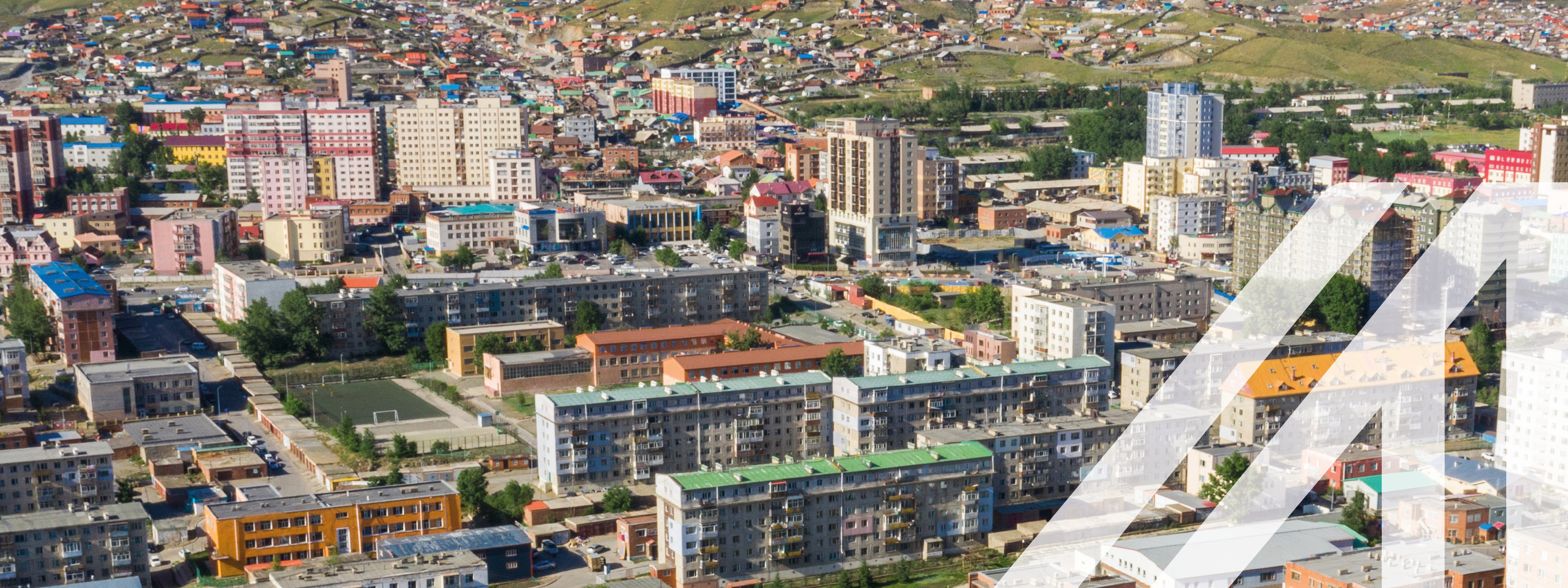 Luftaufnahme von Ulan Bator, Hauptstadt der Mongolei, im Vordergrund stehen viele mehrstöckige Wohnhäuser, im Hintergrund sieht man eine grüne Hügellandschaft.