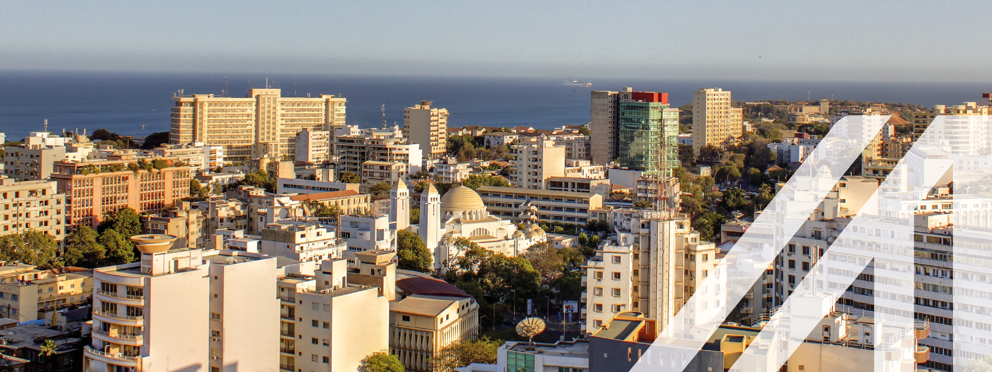 Stadtansicht von Dakar: moderne überwiegend helle Hochhäuser am Meer gelegen unter blauen Himmel. Über das Bild wurde ein weißes Austria A gelegt.