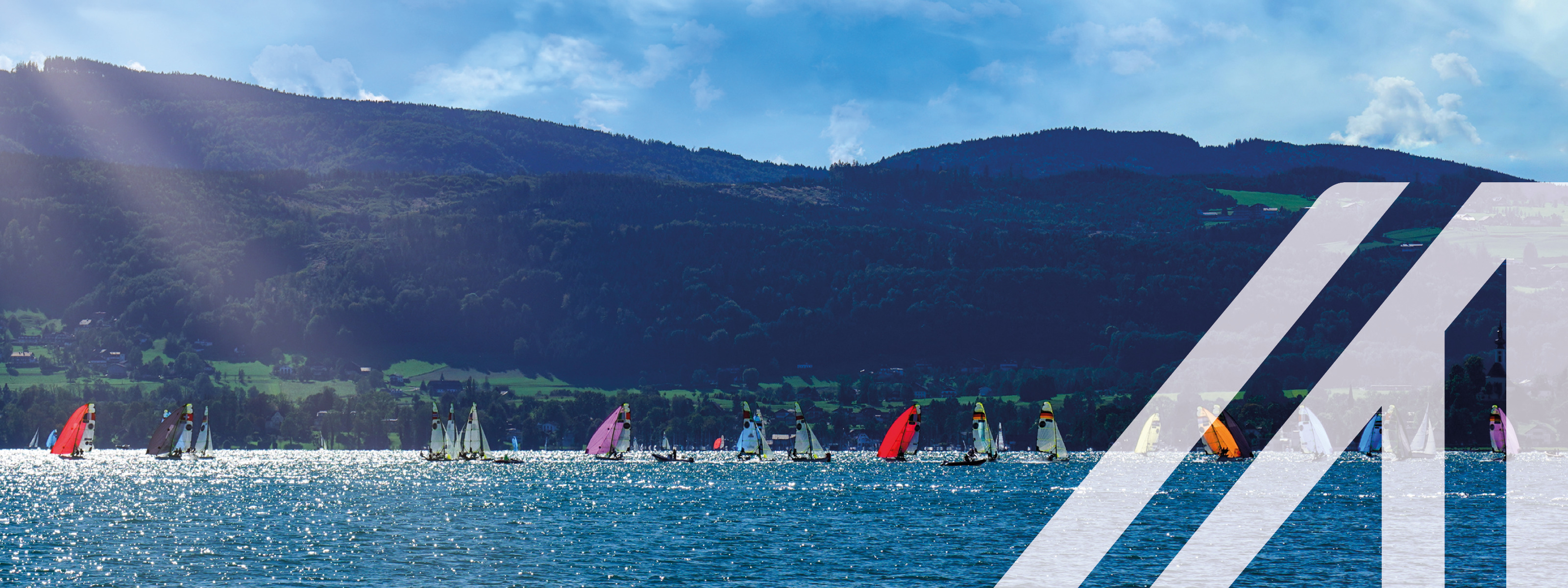 Segelboote mit bunten Segeln bei Weyregg am Attersee bei Sonnenschein unter blauem Himmel, im Hintergrund bewaldete Hügeln und Berge