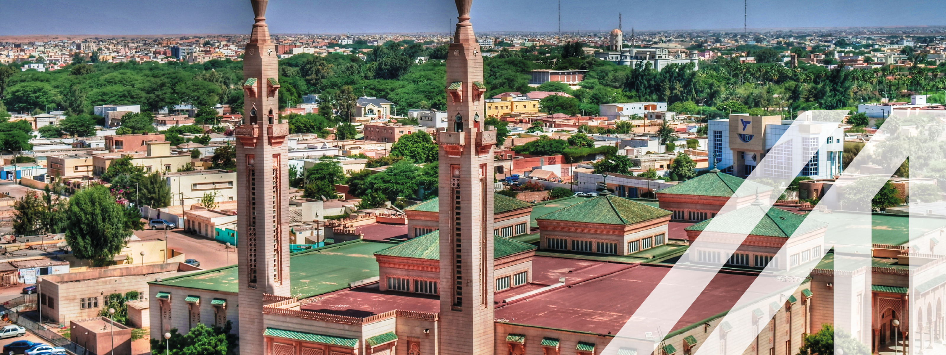 Stadtansicht von Nouakchott: Zentral rosafarbene Moschee mit grünen Dächern und zwei parallel gesetzten Minaretten, im Hintergrund niedrige Häuser und Bäume unter blauem Himmel.