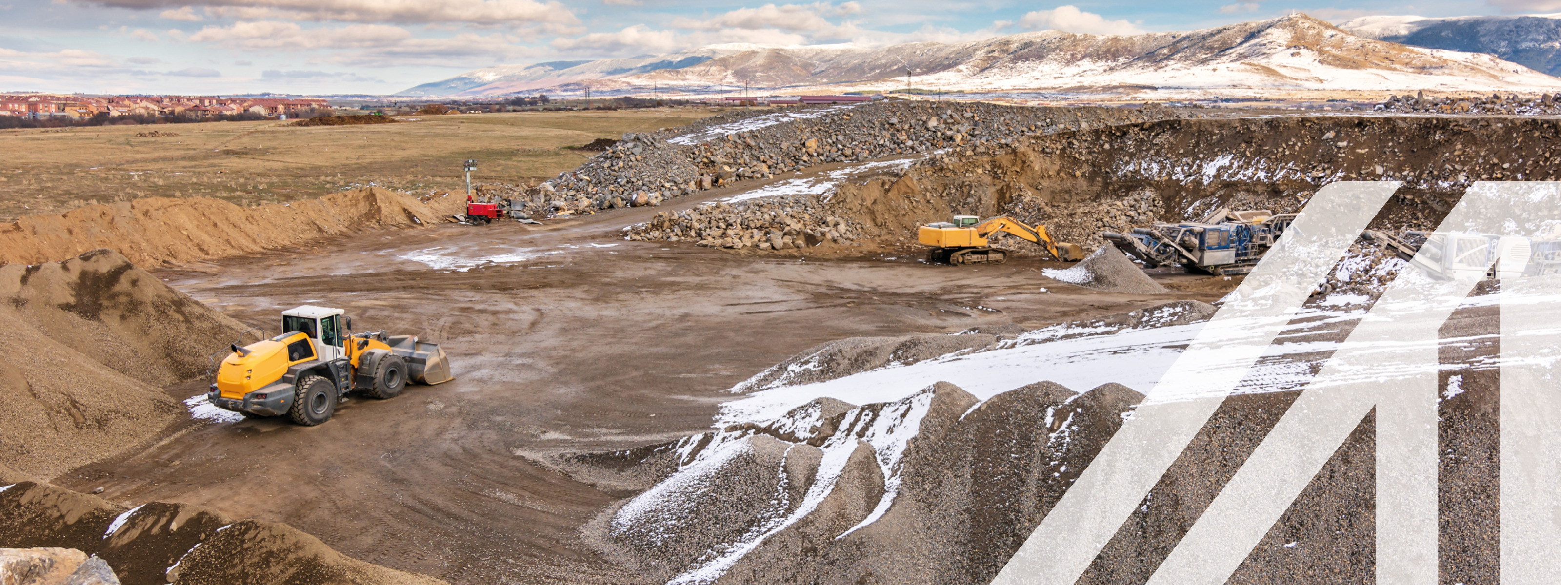 Gelber Bagger, der Gestein in einem teilweise verschneiten Tagebau bewegt, Hügel aus aufgeschüttetem Bergreich, im Hintergrund verschneite Berge und bewölkter Himmel
