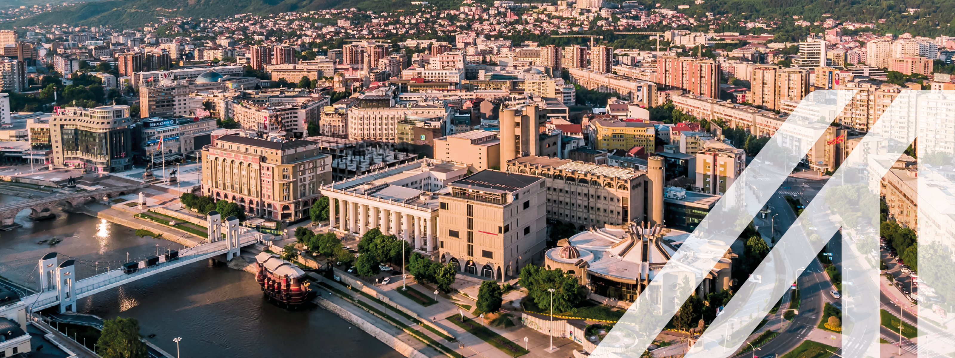 Stadtansicht Skopje: Blick von oben auf die Stadt mit historischen Gebäuden und Monumenten, Fluss Vardar mit Brücken dre Hauptstadt von Mazedonien unter blauem Himmel. Im Hintergrund sieht man eine begrünte hügelige Landschaft, den Berg Vodno.