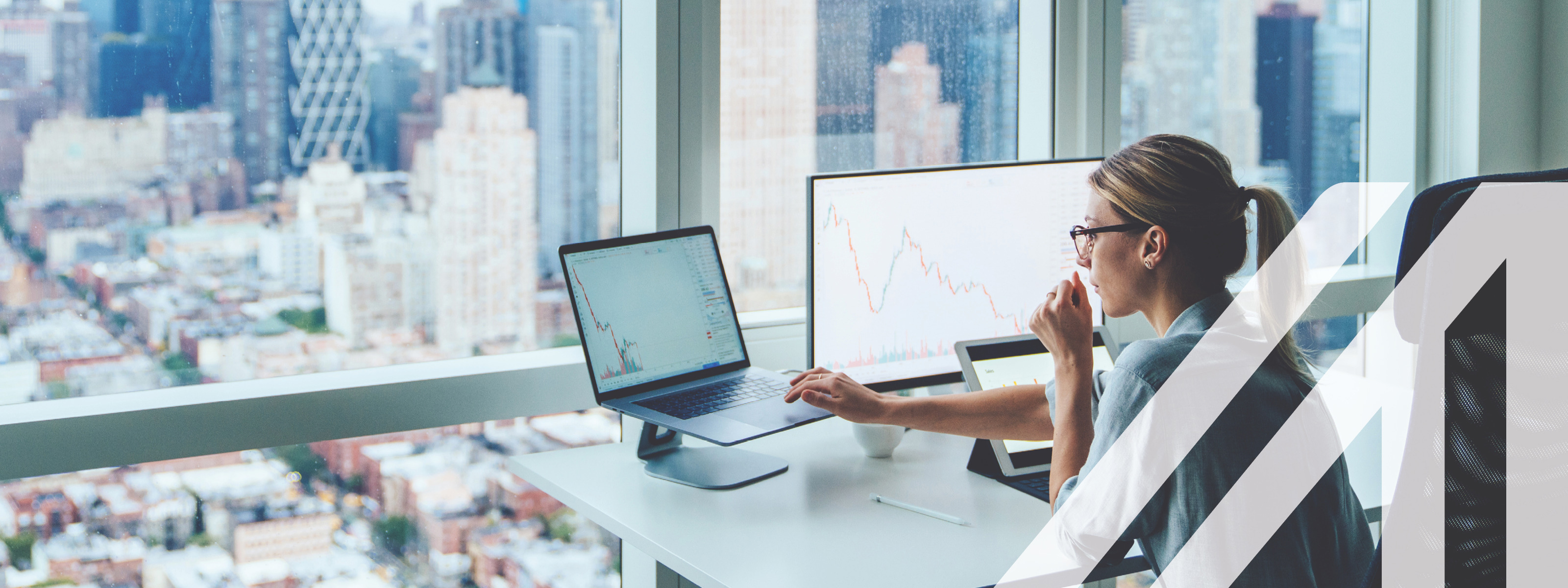Rückansicht einer Business-Frau vor einer Glasfront eines Büros in einem Wolkenkratzer mit Blick auf eine moderne Großstadt. Vor ihr ein Computer mit finanziellen Graphen und Statistiken auf dem Monitor.