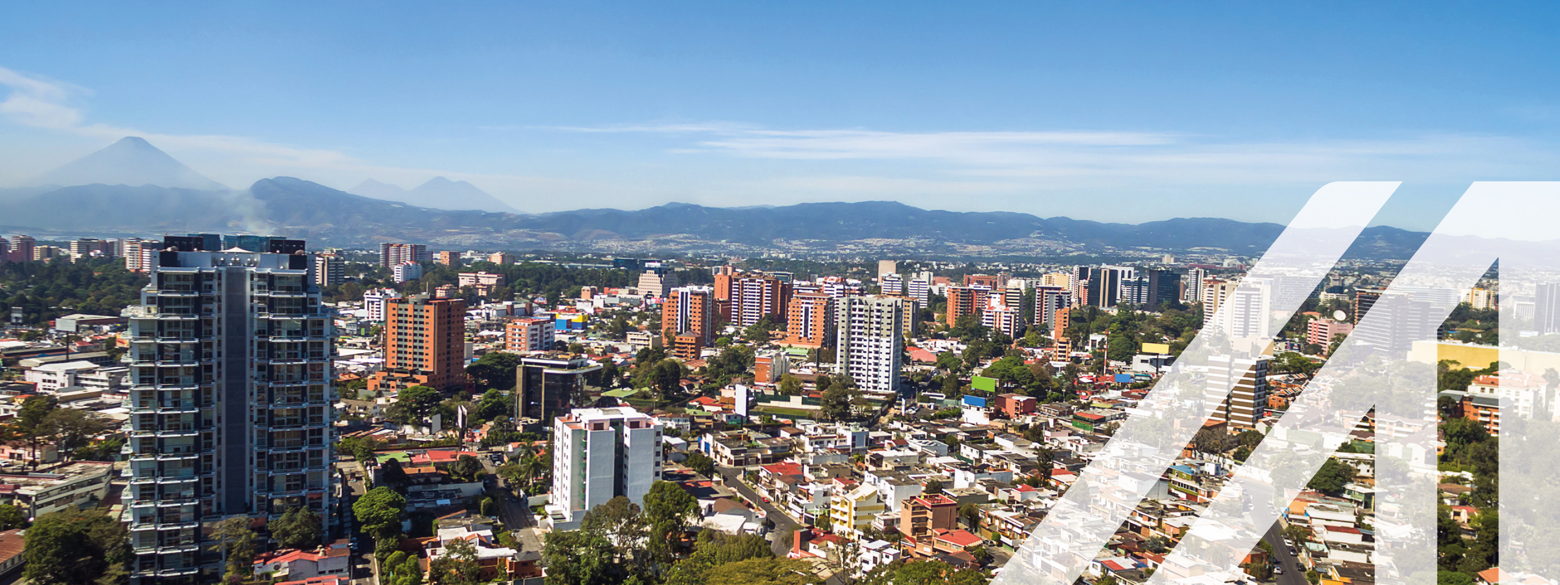 Stadtansicht von Guatemala City: Hochhäuser umgeben von niedrigen Häusern, durchbrochen von grünen Bäumen, im Hintergrund Bergkette unter blauem Himmel