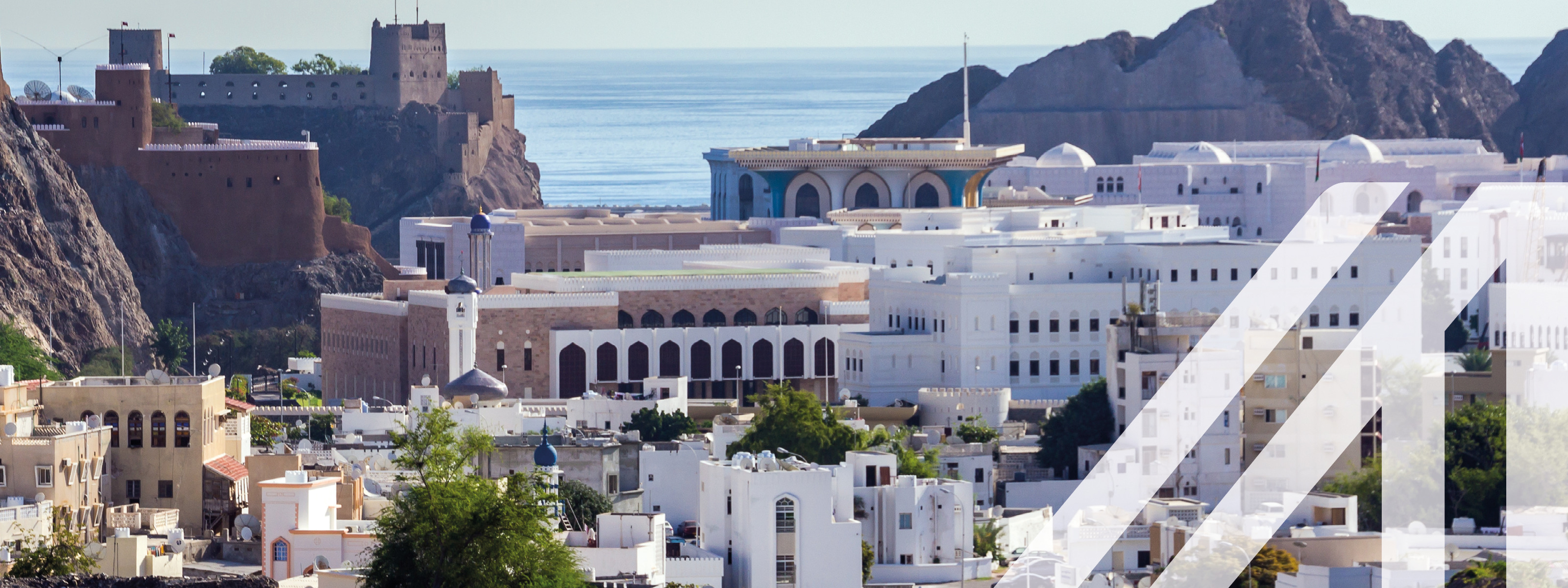 Historische Aldstadt von Maskat mit weißen orientalischen Häusern und einer historischen Festung links im Bild. Im Hintergrund sieht man den Golf von Oman und einen Berg. 
