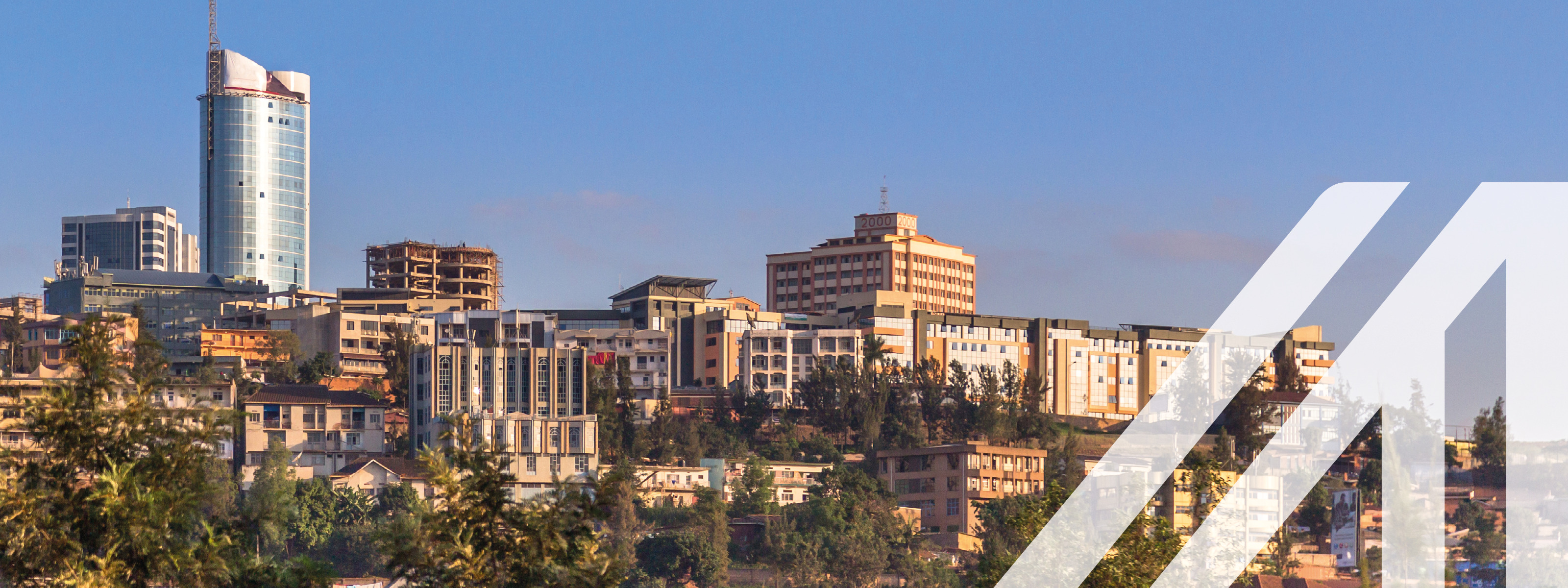 Stadtansicht von Kigali: Im Vordergrund Bäume, im Hintergrund mehrstöckige Häuser auf Hügel gelegen, darüber blauer Himmel