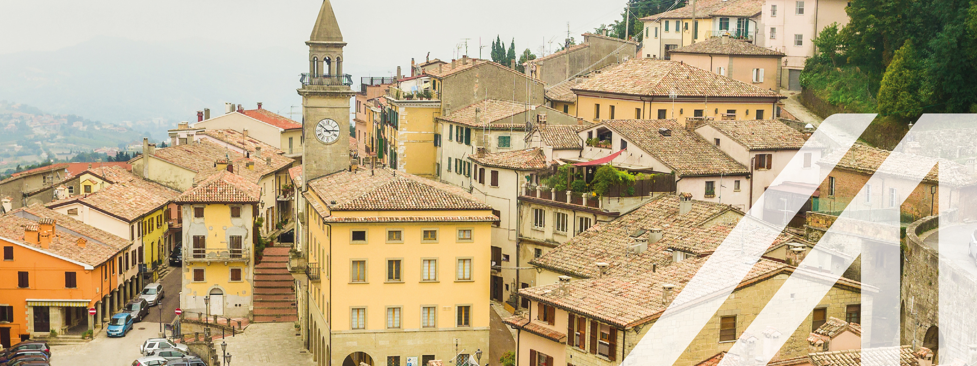 Stadtansicht von San Marino: Vorwiegend gelbbeige Häuser auf Hügel verlaufend mit Schindeldächern, Uhrturm hervorragend, im Zentrum Platz mit parkenden Autos, daneben Straße und grüner Laubbaumwald