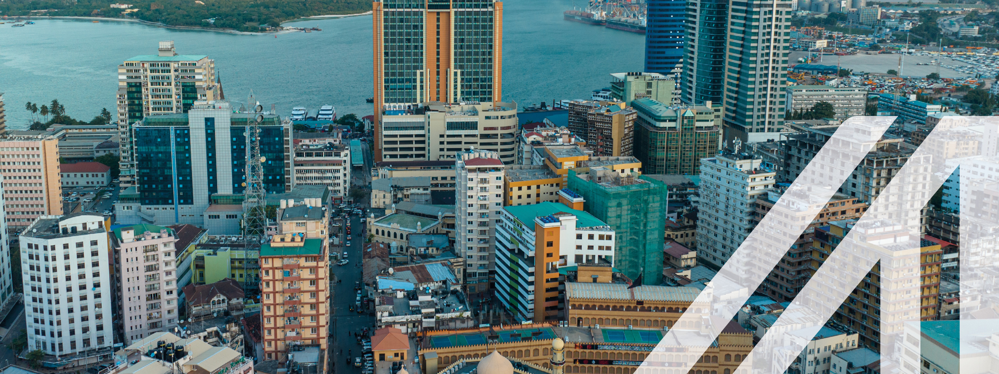 Luftaufnahme von Dar Es Salaam in Tansania: bunte Hochhäuser unter blauem leicht bewölkten Himmel, im Hintergrund erkennt man den Ozean. Über das Bild wurde ein weißes Austria A gelegt.<br />
