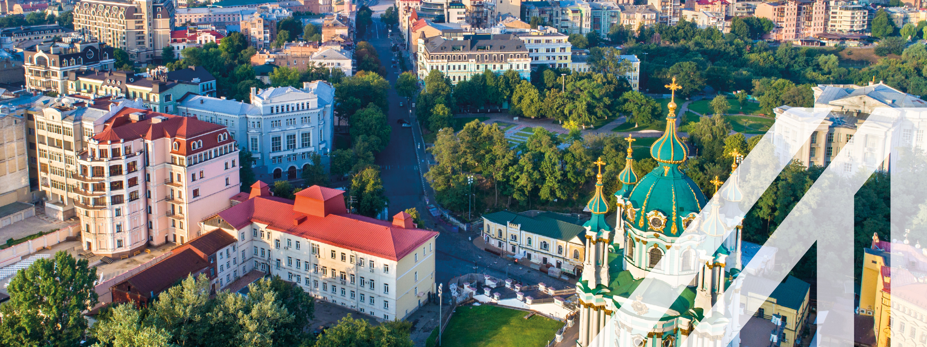 Stadtansicht von Kiew: eine breite Straße zieht sich durch die Stadt mit der Sankt-Andreas-Kirche und modernen Gebäuden, aufgelockert durch viele Bäume. Über das Bild wurde ein weißes Austria A gelegt.<br />
