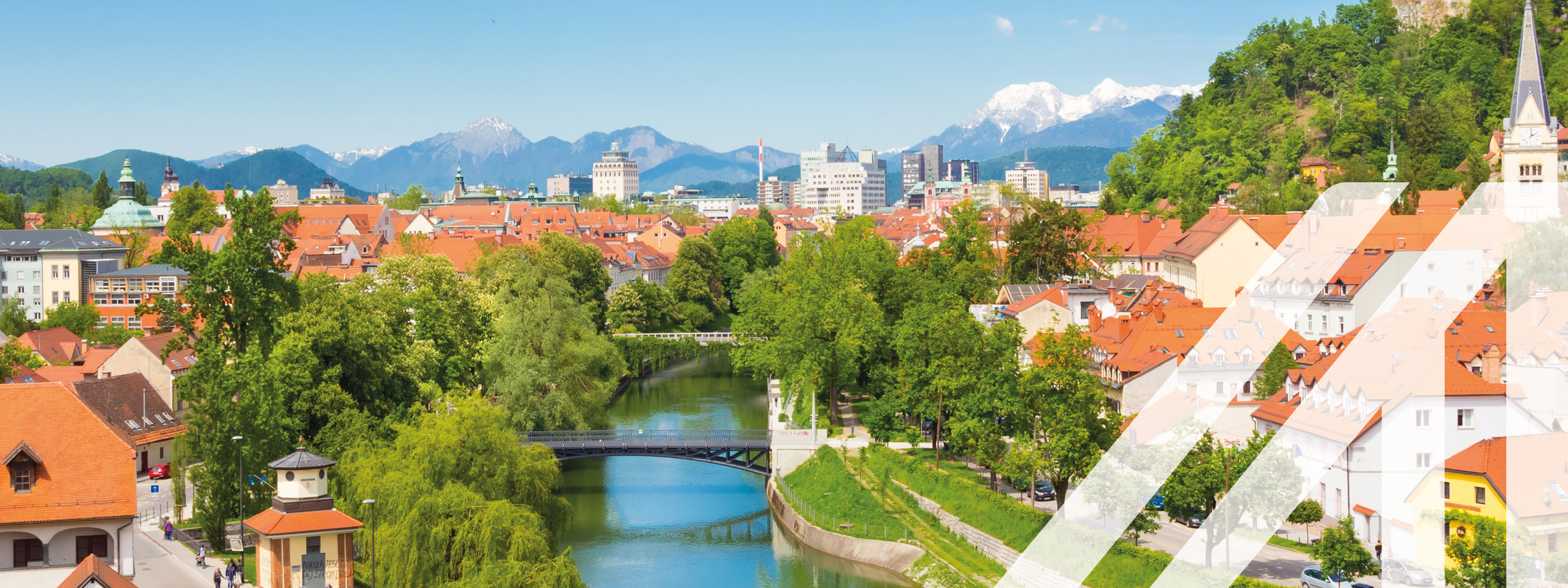 Blick auf Sloweniens Hauptstadt Laibach mit vielem weißen Häusern und roten Dächern. In der Mitte schlängelt sich ein Fluss mit Brücken durch die Stadt, rechts im Bild sieht man auf einer Anhöhe eine Burg, im Hintergrund verschneite Berggipfel. 