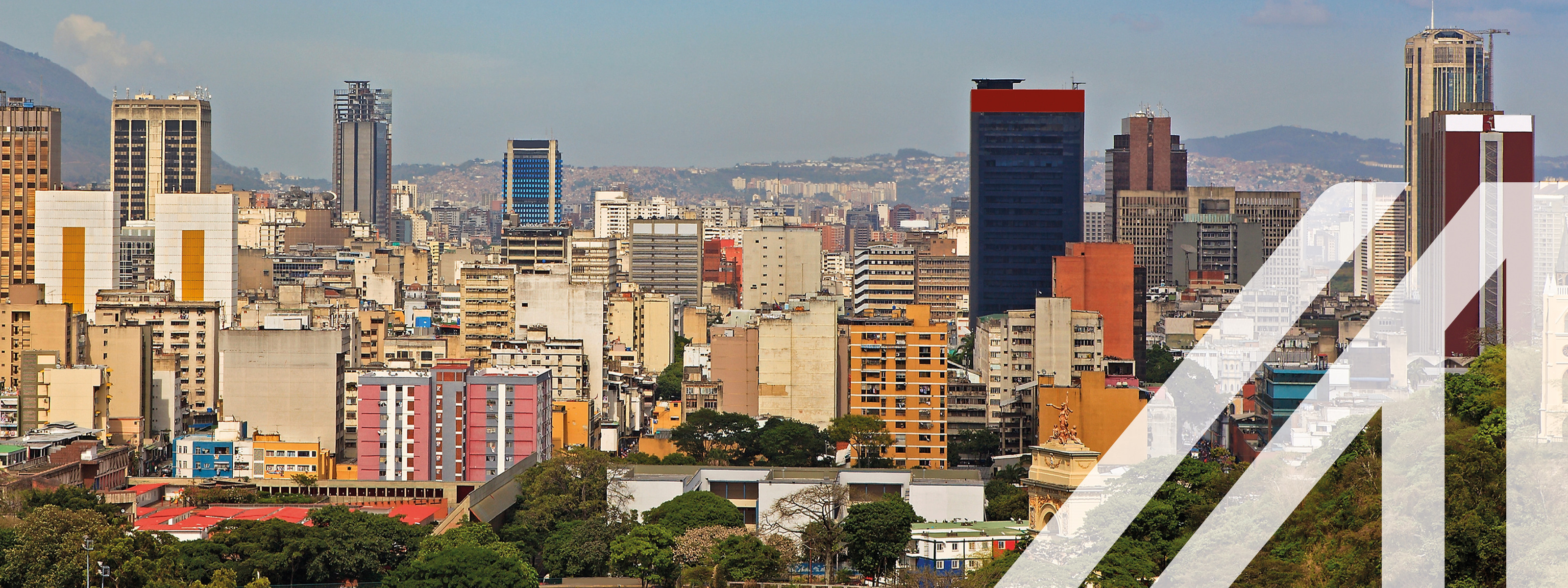 Stadtansicht von Caracas: Skyline mit zahlreichen Wolkenkratzern in Downtown Caracas, Hauptstadt von Venezuela, unter blauem Himmel, gelegen vor einer Bergkette. Über das Bild wurde ein weißes Austria A gelegt.