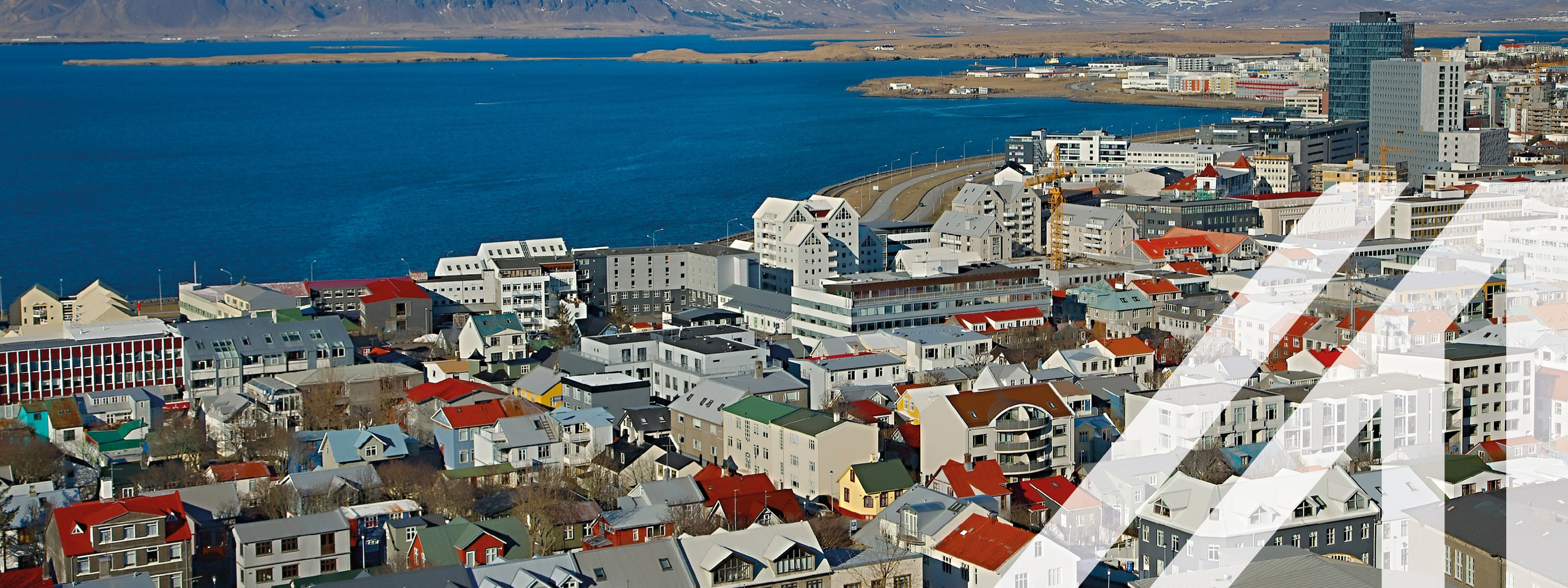Stadtansicht von Reykjavik mit vielen roten Dächern mit modernen Hochhäusern; im Hintergrund sieht man das Meer und verschneite Berge