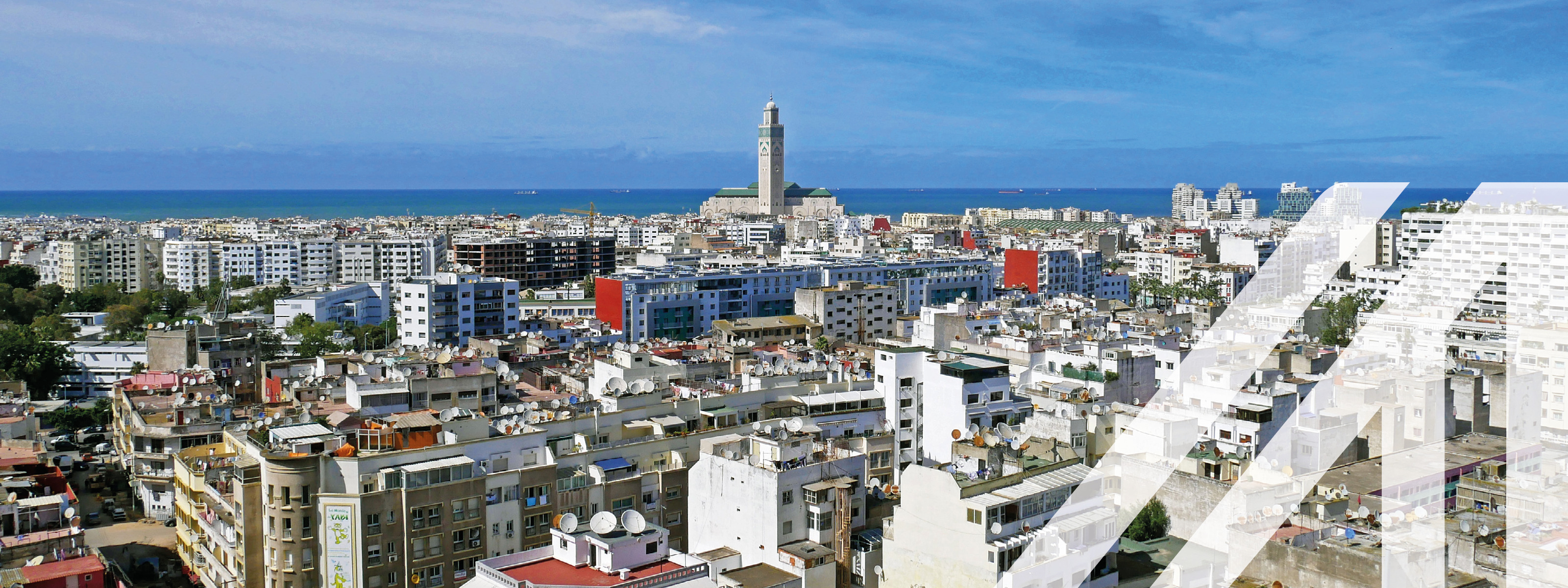 Panoramablick auf Casablanca, der Turm der Hassan II Moschee ragt hervor, unter blauem Himmel am Meer gelegen.

