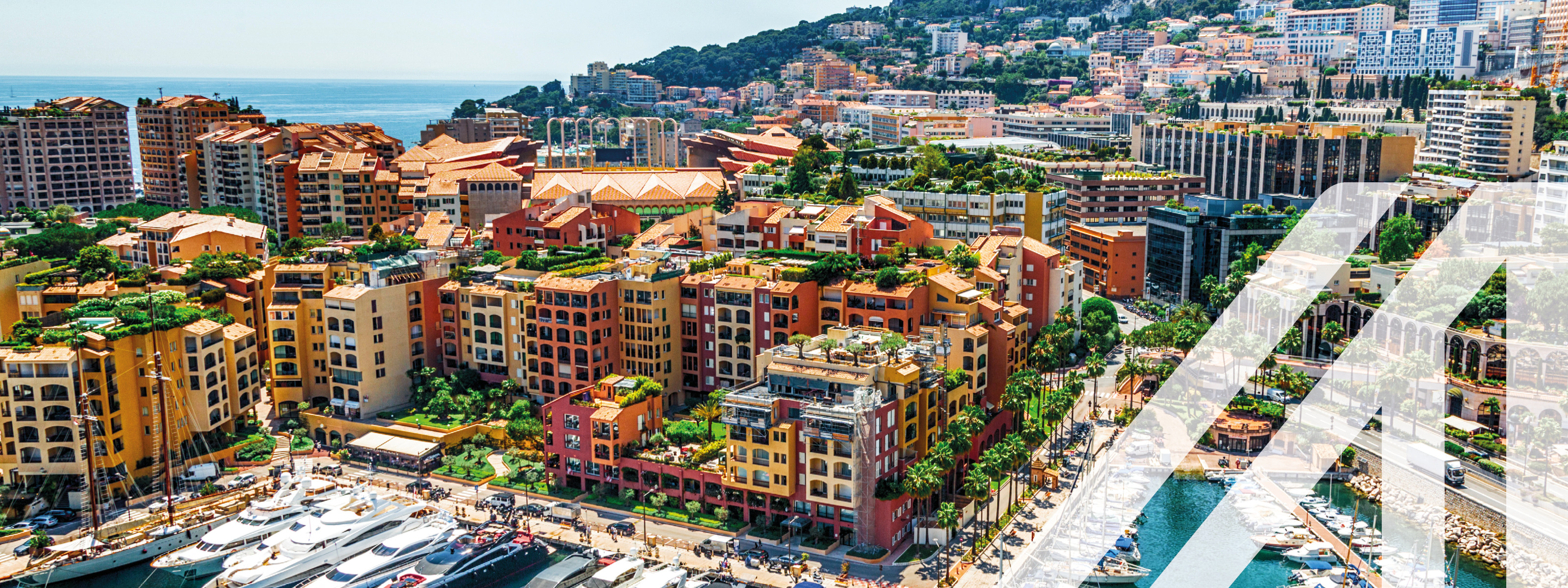 Blick auf den Hafen von Monte Carlo mit bunten Hochhäusern und Yachten, im Hintergrund sieht man das offene Meer und eine ansteigende bewaldete Bergkette  