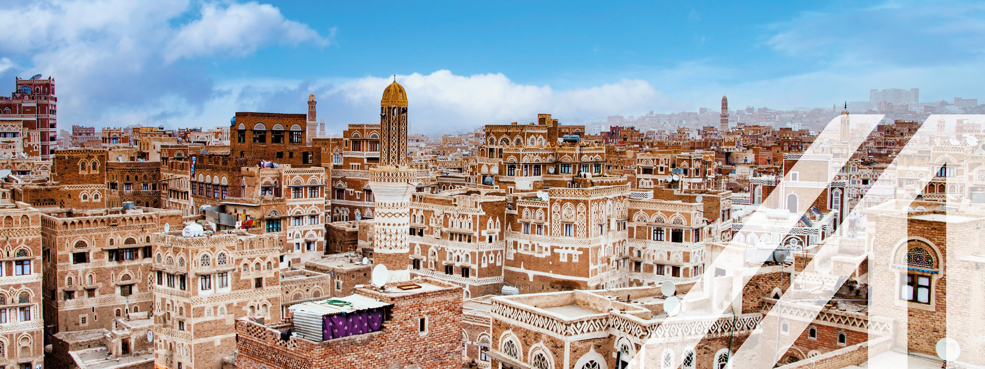 Stadtansicht von Sanaa: Die Straßen der Altstadt von Sanaa, der Hauptstadt des Jemen. Terrakottafarbene Häuser mit weißen Verzierungen unter blauem Himmel, in der Mitte ein Turm. Über das Bild wurde ein weißes Austria A gelegt.
