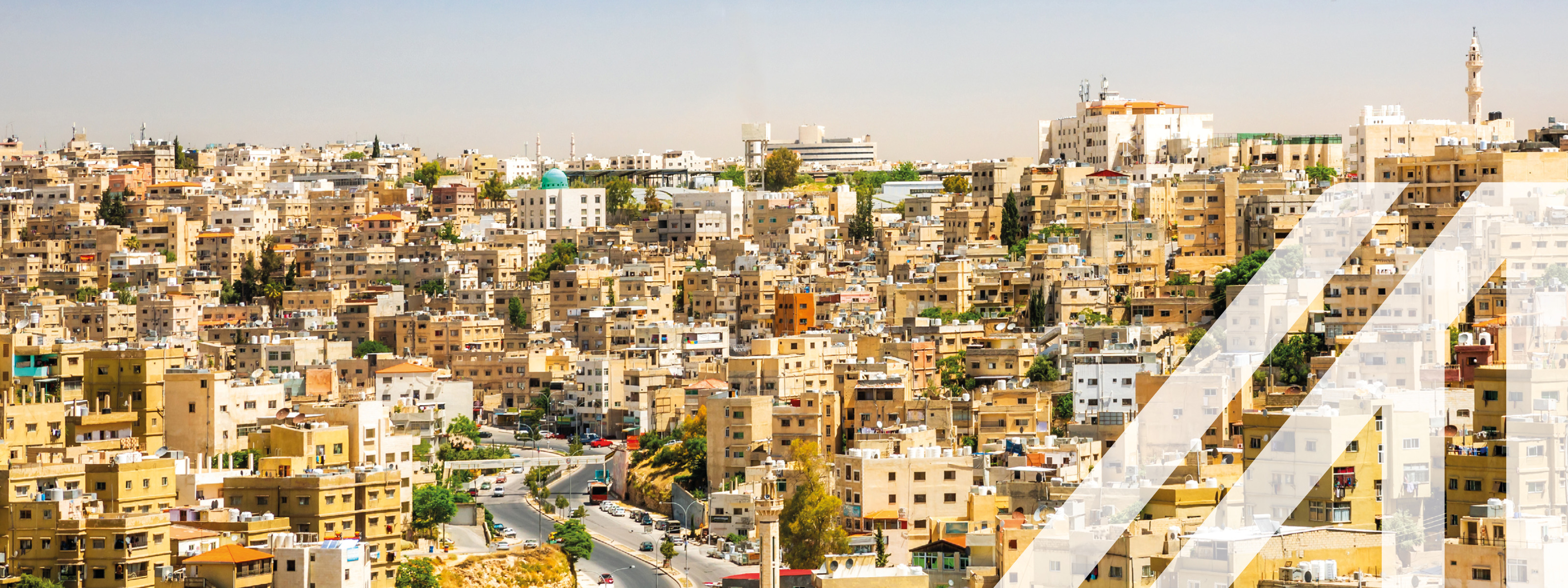 Stadtansicht von Amman: Blick auf viele helle Häuser unter blauem Himmel, eine Straße schlängelt sich bergauf. Über das Bild wurde ein weißes Austria A gelegt.