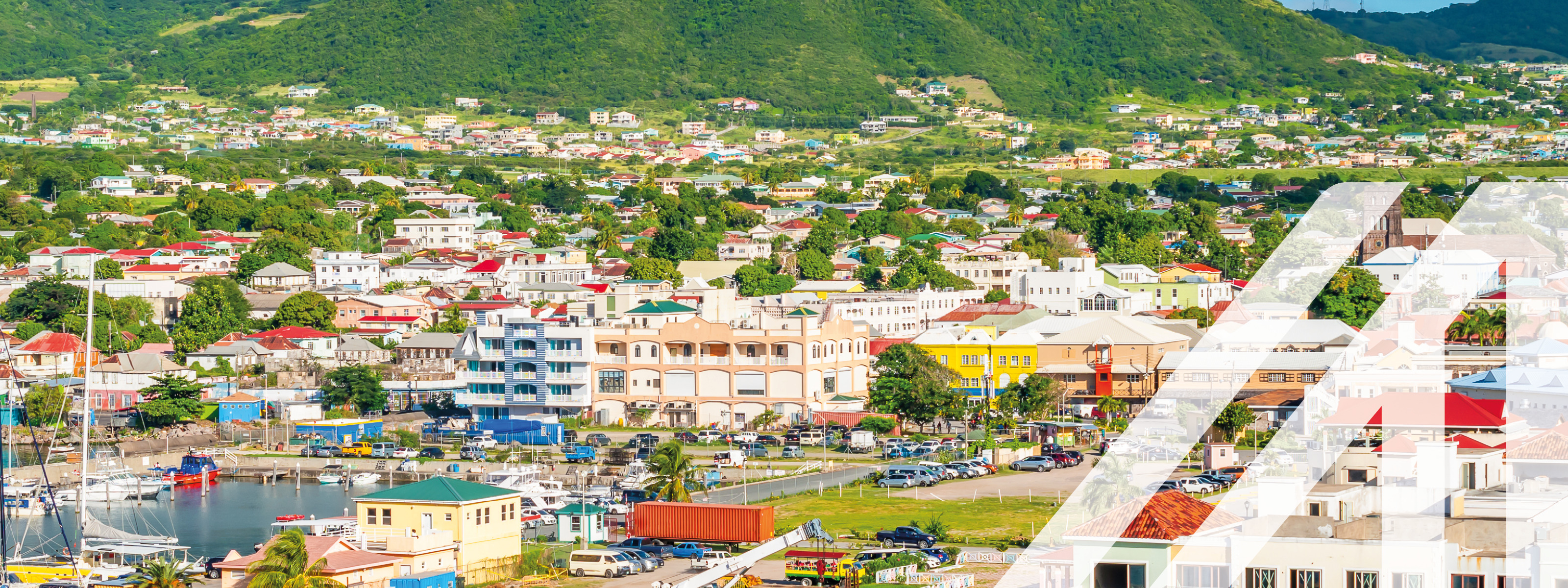 Ansicht von Basseterre: im Vordergrund Wasser und Hafen sowie kleine Stadtlandschaft, im Hintergrund begrünte Hügel unter wolkenverhangenem Himmel