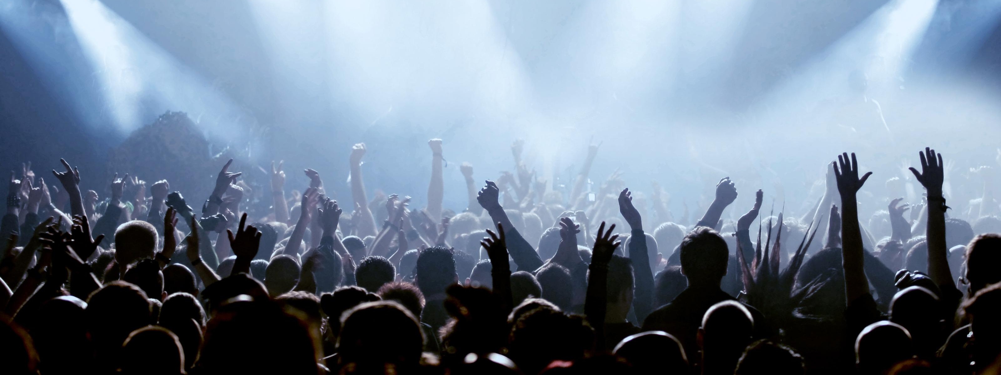 Viele Personen, Fans, stehen vor einer Bühne, die mit Scheinwerfern beleuchtet ist