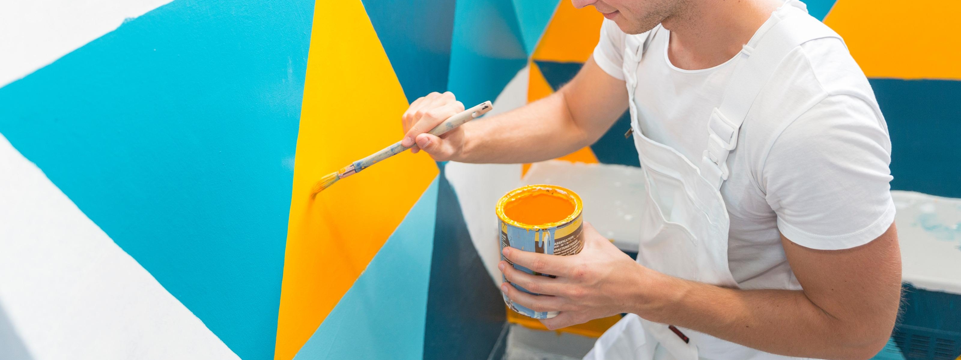 Person in weißem Arbeitsgewand streicht gelbe Farbe an Wand, die mit blauen, gelben und türkisen Dreiecken gemustert ist