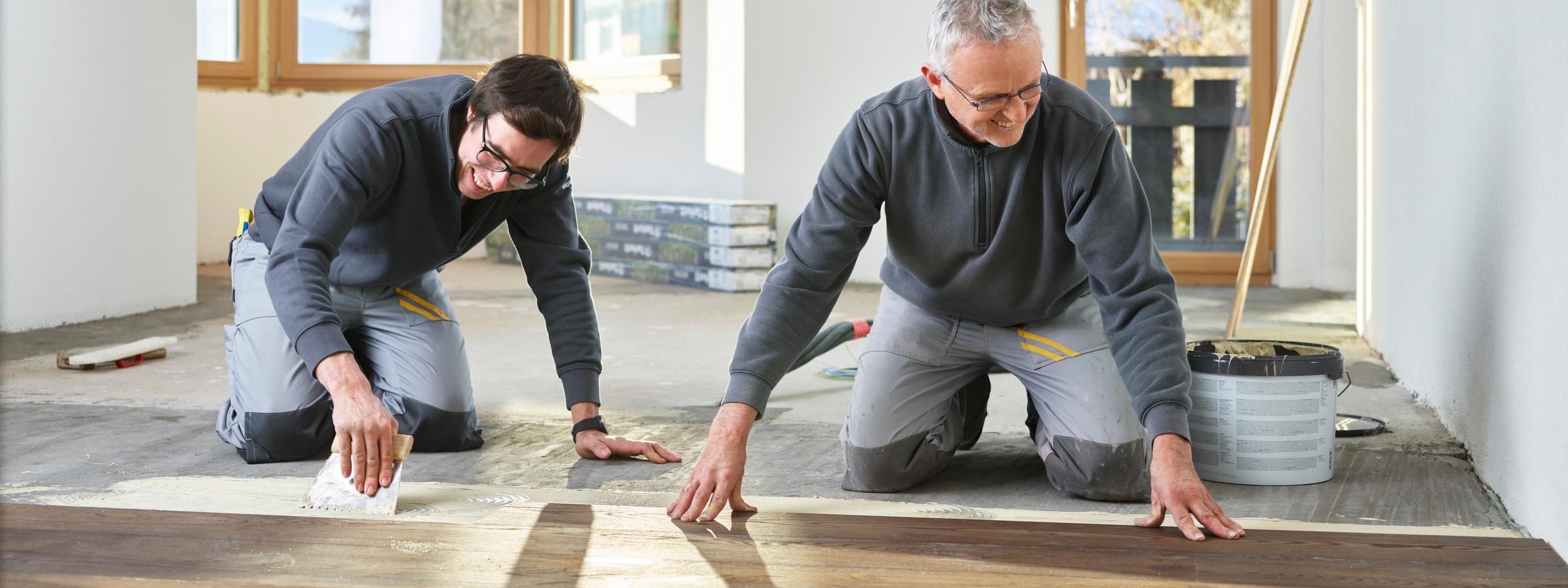 Zwei Personen in grauen Arbeitsmonturen knien am Boden und verlegen Holzbodenbretter, im Hintergrund Fenster, Balkontüre und Fernseher