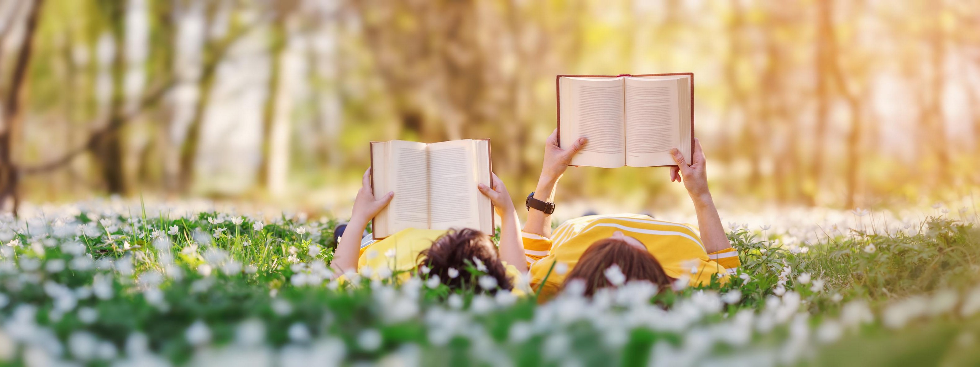 Zwei Personen liegen in Blumenwiese und lesen Bücher, im Hintergrund Wald