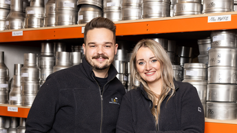 Lukas Steiner und Lisa Hupfauf stehen gemeinsam vor Regalen mit Edelstahlteilen in einem Lager. Beide tragen dunkle Firmenjacken mit EAH-Logo und lächeln in die Kamera. Im Hintergrund sind zahlreiche Metallteile und beschriftete Regalböden zu sehen.