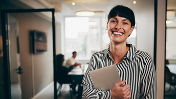 Porträt einer lächelnden Person mit dunklen Stirnfransen und gestreifter Bluse, die in einem Flur steht und mit beiden Händen einen Laptop beim Körper hält, im Hintergrund zeigen sich helle Meetingräume