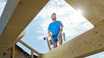 Blick von unten auf eine Person mit blauem T-Shirt, die auf Leiter steht und zwischen Holzbalken hervorlugt 