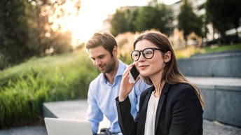 Person mit langen dunklen Haaren, Businesskleidung und Brille sitzt auf einer Stufe und telefoniert während eine andere Person daneben mit einem Laptop arbeitet
