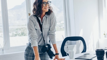 Lächelnde Person mit umgehängter Fotokamera über Tisch mit Fotos lehnend, ringsum verschiedene Utensilien