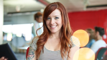 Person mit rötlichen langen Haaren trägt mit gestreiftem Shirt, Tattoo am rechten Oberarm blickt freudig in die Kamera und hält ein Tablet in der Hand, im Hintergrund  sitzen und stehen weitere Personen in einem roten Raum