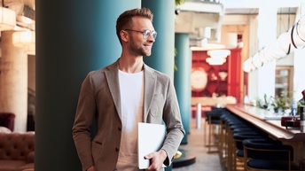 Lächelnde Person mit Brillen hält Notebook in der Hand und steht vor blauer Säule in einem Lokal, im Hintergrund Bar mit Hockern