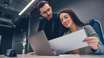 Person in Businesskleidung sitzt an einem Schreibtisch mit Laptop und hält freudig ein Dokument mit einer Grafik in der Hand während eine weitere Person interessiert daneben steht und darauf blickt