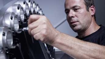 Eine Person mit kurzen, braunen Haaren und schwarzem Oberteil hantiert mit einem Werkzeug an einem Objekt aus Metall mit runden Einfassungen